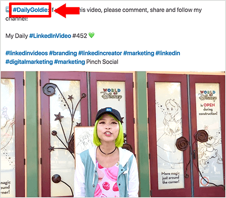 Aceasta este o captură de ecran care ilustrează modul în care Goldie Chan folosește hashtaguri în textul postărilor sale video pe LinkedIn. Înștiințările roșii indică hashtagul #DailyGoldie din text, care este unic pentru postările sale video și o ajută să urmărească partajările. Postarea include, de asemenea, alte hashtag-uri relevante care îi ajută pe oameni să-i găsească videoclipul, inclusiv #LinkedInVideo. În imaginea video, Goldie stă în fața unor uși la un ecran World of Disney. Este o femeie asiatică cu părul verde. Poartă o șapcă neagră LinkedIn, un colier choker negru, o cămașă roz cu imprimeu macaron și o jachetă albastră și albă.